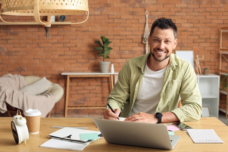 homem empreendedor sorrindo para a câmera enquanto trabalha em seu notebook, superando os desafios do empreendedorismo