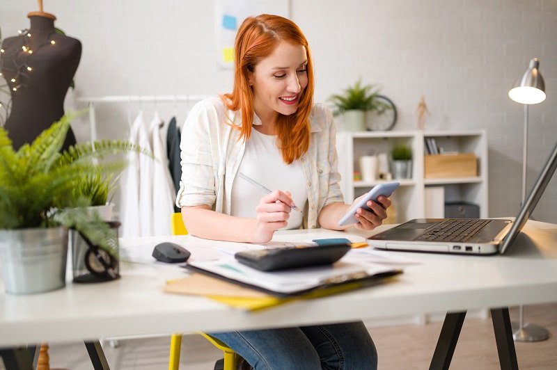 mulher sorridente trabalhando em seu ateliê na mesa com computador, anotando e verificando informações sobre seu negócio