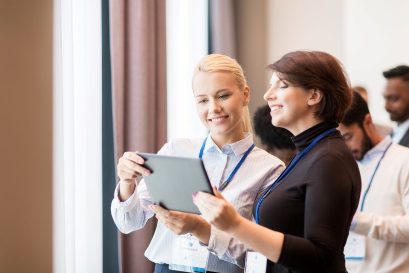 Duas mulheres conversam utilizando um tablet em um escritório com outros profissionais ao fundo. 
