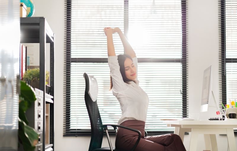 Mulher, em sua rotina de trabalho, sentada em frente ao computador, alonga os braços e as costas.