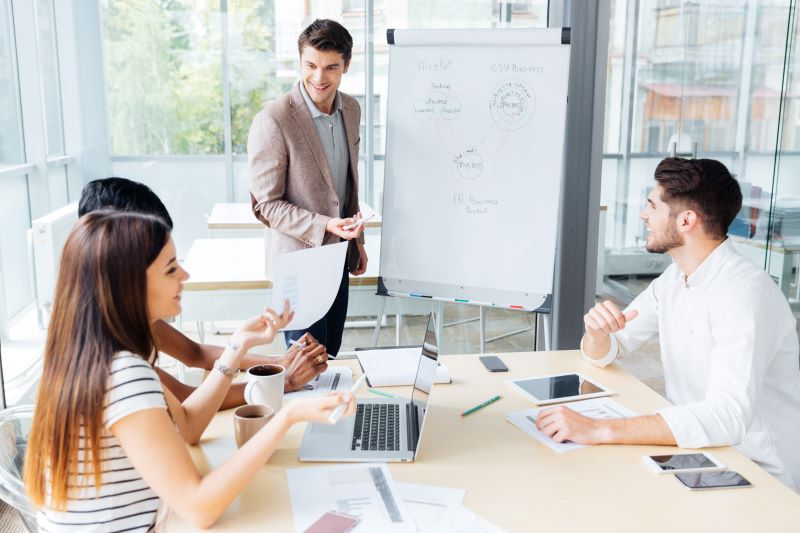 homem explica conceitos de tomada de decisão em quadro branco para seus três colegas sentados em mesa de reuniões