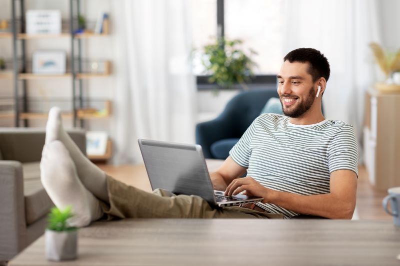 homem sorridente trabalhando em sua casa, com roupas casuais e sem os sapatos, apoiando os pés sobre a mesa com o computador no colo
