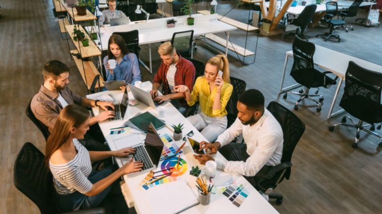 Coworking em são paulo​: várias pessoas reunidas em mesa de coworking com computadores e elementos gráficos.