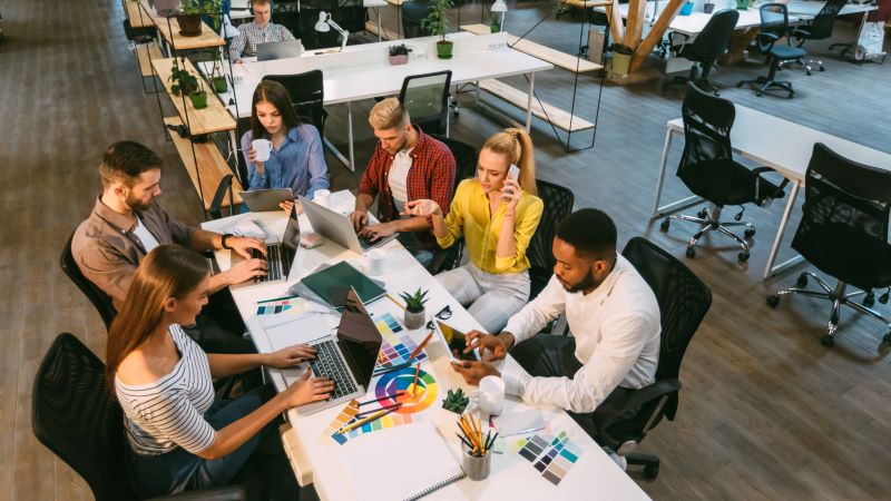 Coworking em são paulo​: várias pessoas reunidas em mesa de coworking com computadores e elementos gráficos.