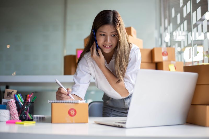 Pequenos negócios que dão lucro​​: empreendedora preparando produto para envio enquanto fala ao telefone.