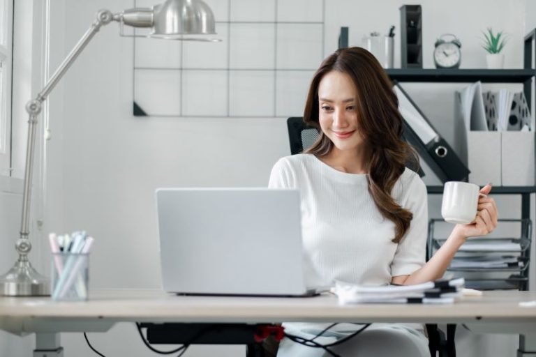 trabalho híbrido​: mulher sorridente trabalhando no escritório enquanto toma café
