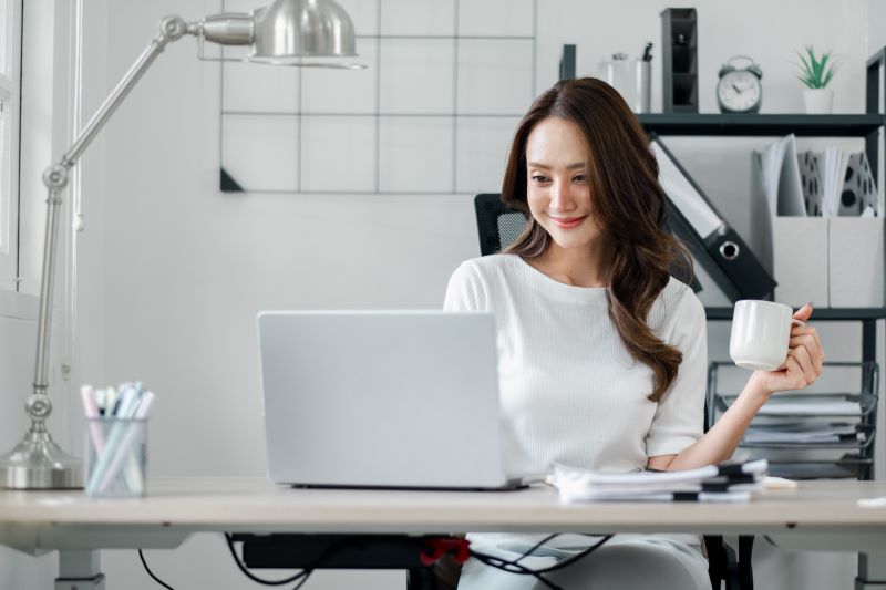trabalho híbrido​: mulher sorridente trabalhando no escritório enquanto toma café